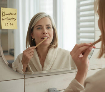 Kvinna i 50-årsåldern borstar tänderna med bambutandborste i badrum, med post-it-lapp på spegeln om hormonläkare – illustration av munhälsa i klimakteriet.